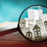 Magnifying glass in front of an open newspaper with paper houses. Concept of rent, search, purchase real estate.