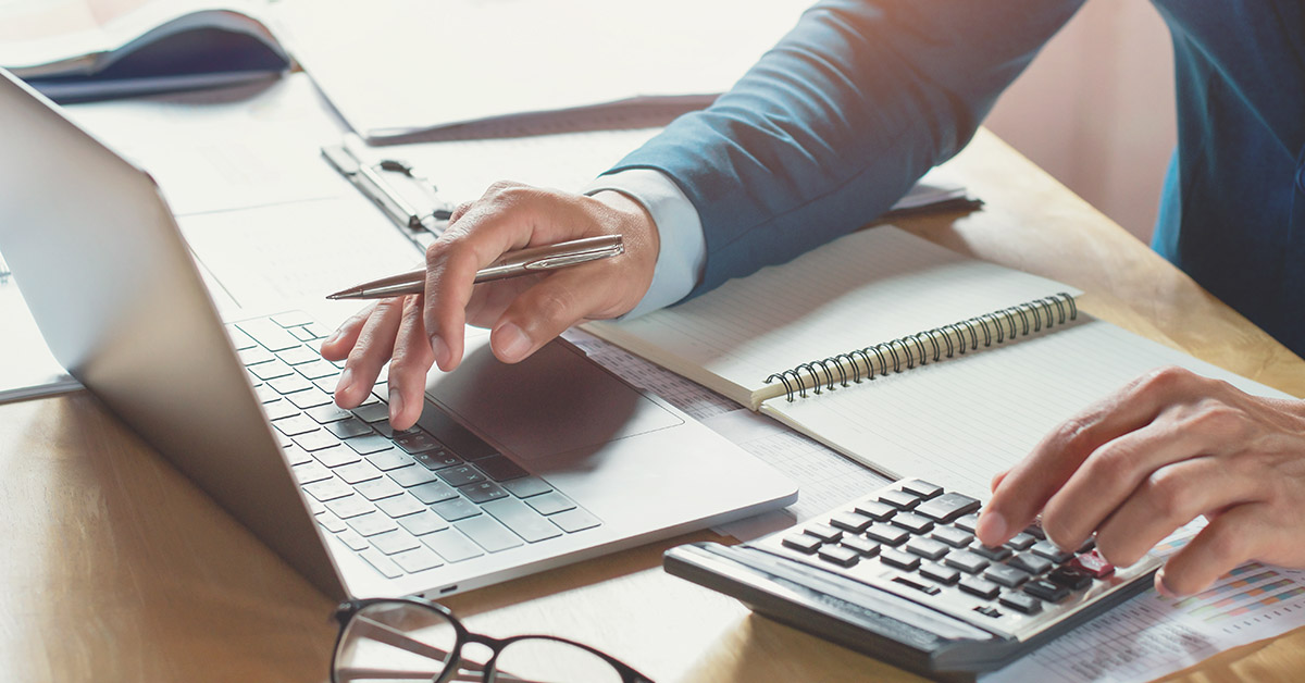 Tax attorney multitasking at a desk, using a laptop and calculator, surrounded by documents and a notepad, illustrating 501(c)(3) tax obligations.